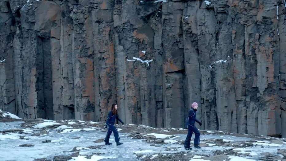 snowy ground against a rock cliff with two people walking